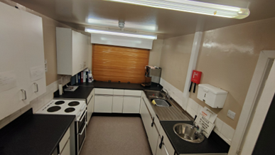  A small, functional kitchen with white cabinets, dark countertops, a stove on the left, and a sink on the right, all illuminated by fluorescent lights.
