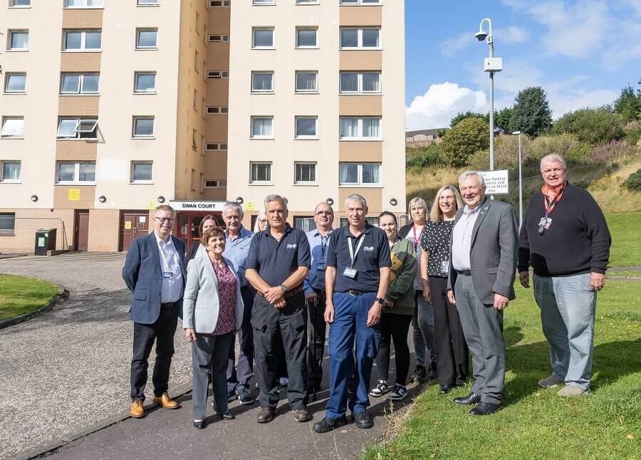 Group of people standing in front of a residential building labelled 'Swan Court' on a sunny day - Mitigation measures as work on Methil tower blocks progresses
