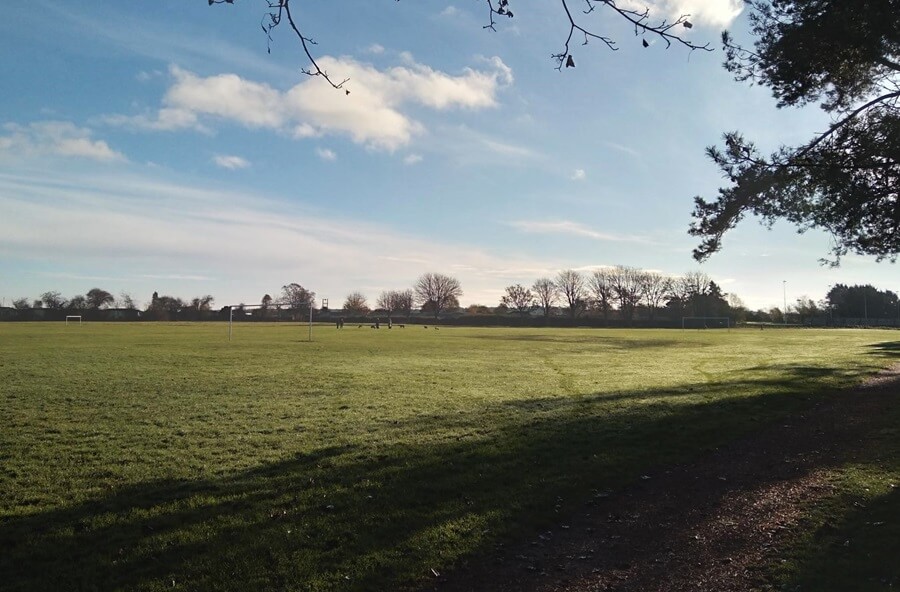 Large grassy sports field with goalposts in the distance, bordered by trees and a dirt path. Clear blue sky with scattered clouds and long shadows on the grass.