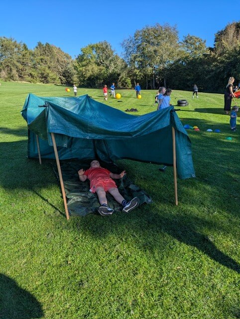 Child lying under a green tarp shelter on a sunny grassy hill.