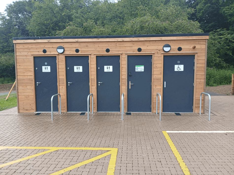 Modern wooden restroom building with three unisex doors and one accessible door, set in a park with trees in the background.
