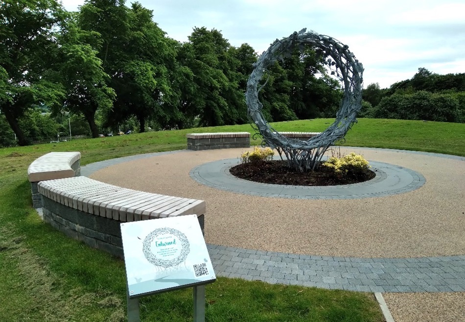 Metal ring sculpture in a landscaped area with benches and a sign commemorating community contributions