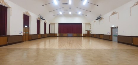 An empty assembly hall with a polished wood floor, white walls, and a stage with red curtains at the far end.