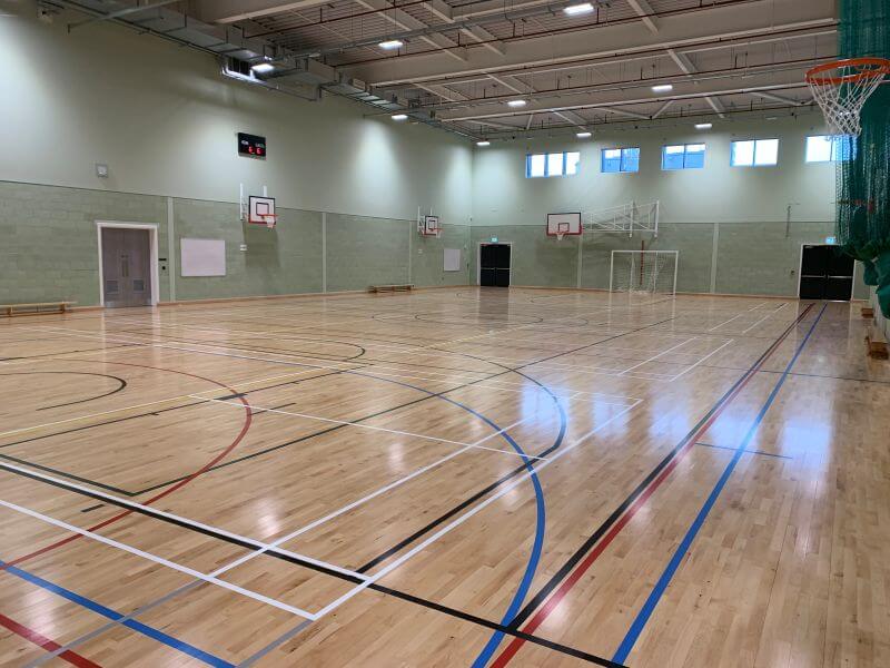 A bright indoor sports hall with a high wooden ceiling and court markings on a polished floor. A basketball hoop with a backboard is mounted on the right wall. Near the hoop, two players are jumping toward the basket while others move across the court in the background. The hall is spacious, with pale walls, green lower panels, and clear lines for multiple indoor sports.