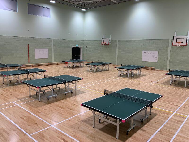 Indoor games hall with a polished wooden floor marked for court sports, high pale-green walls, and two wall-mounted basketball hoops at either end.