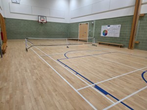 An indoor gymnasium with a polished light-wood floor and a portable sports net set up across the center of a court marked with white and blue lines. The lower half of the walls consists of green brick, while the upper half is white. A basketball hoop is mounted on the far wall, and a colorful poster hangs near a double wooden door.