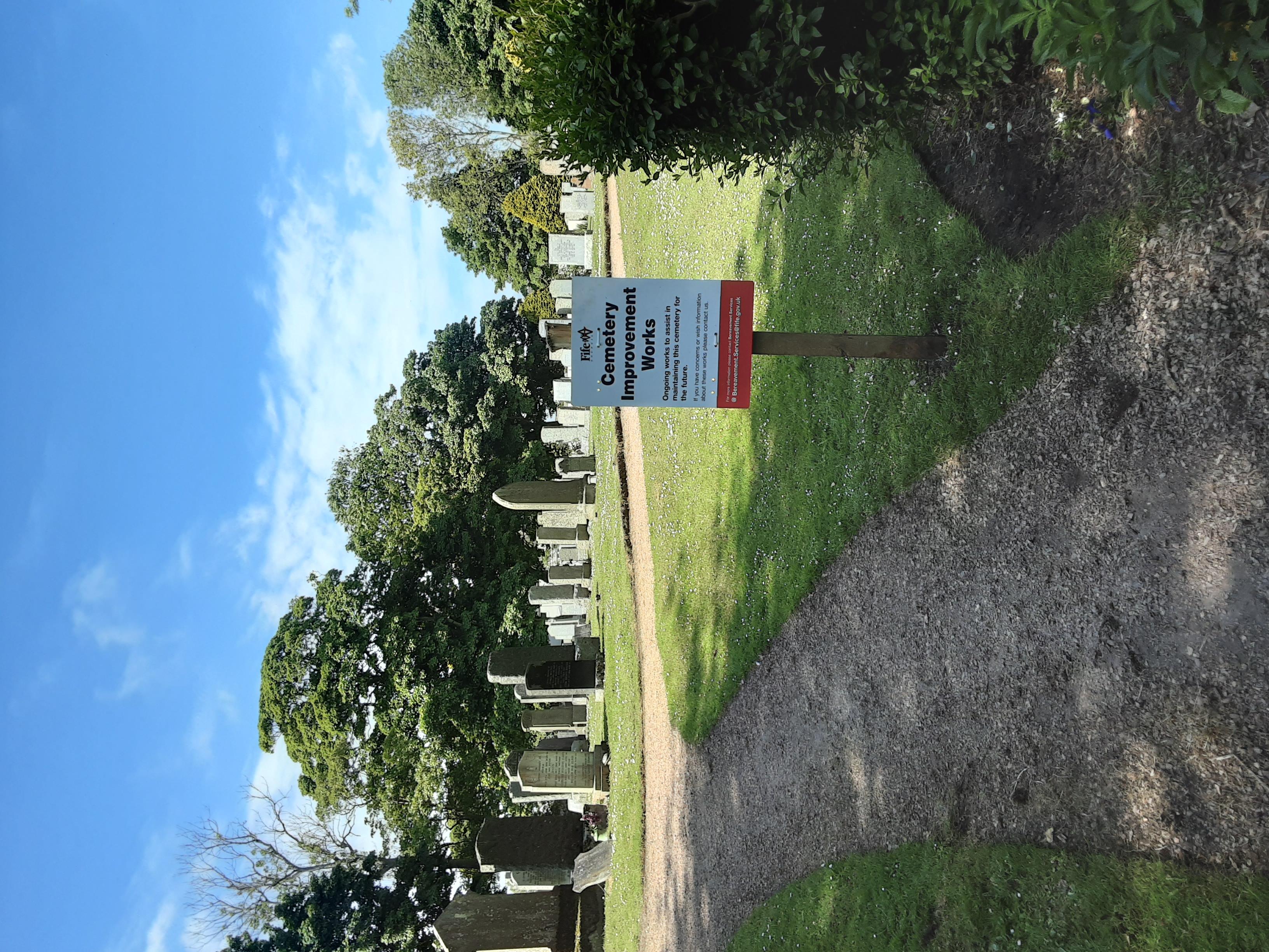 Entrance to Crail Cemetery on a sunny day with headstones visible in the background.   In the foreground there's a sign advising that headstone inspections are taking place .