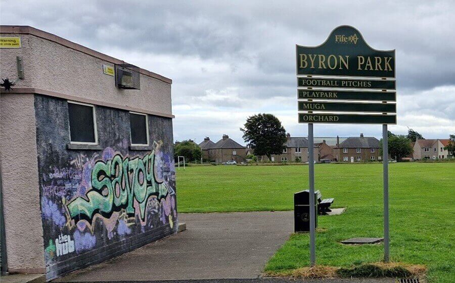The Savoy park sign A sign says Byron Park. The Savoy building with graffiti stands left. Green fields and houses in the background