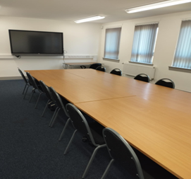 meeting room a meeting room with a long, light-colored table surrounded by black chairs. A large television screen is mounted on the white wall at one end of the table. The floor is covered with a dark blue carpet. There are windows with blinds on the left side of the room and a doorway on the right.