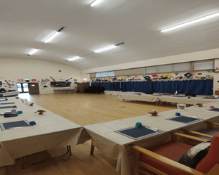 A wide shot of a community hall with light wood flooring. Several rectangular tables, covered with white tablecloths and set with place settings, are arranged in a U-shape. Blue curtains hang on the far wall, decorated with colorful paper shapes.