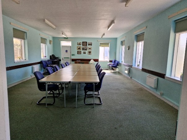 A conference or meeting room with light blue walls and several windows. There is a long rectangular table in the center of the room with multiple blue chairs arranged around it. Additional stacked chairs are visible in the back left corner of the room. The walls have several framed pictures, and there is a door at the far end of the room. The floor is carpeted, and fluorescent lights are mounted on the ceiling.