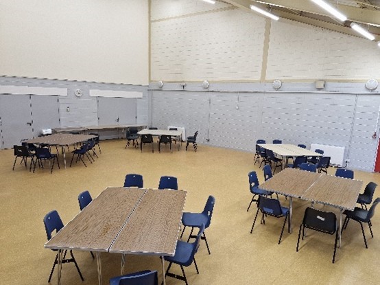 A large room with multiple tables and chairs arranged in groups. The tables are rectangular, and the chairs are blue. The room has a high ceiling with fluorescent lights and grey walls.