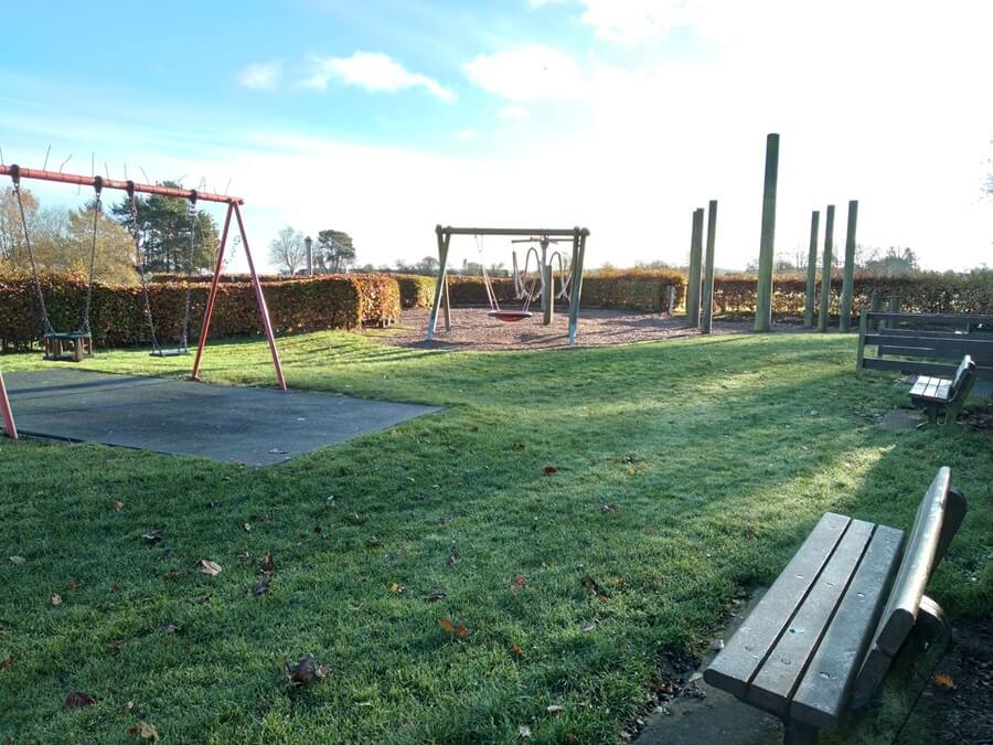 Playground with swings and tall wooden poles. Surrounded by grass, few benches, trees and hedges under a partly cloudy sky.
