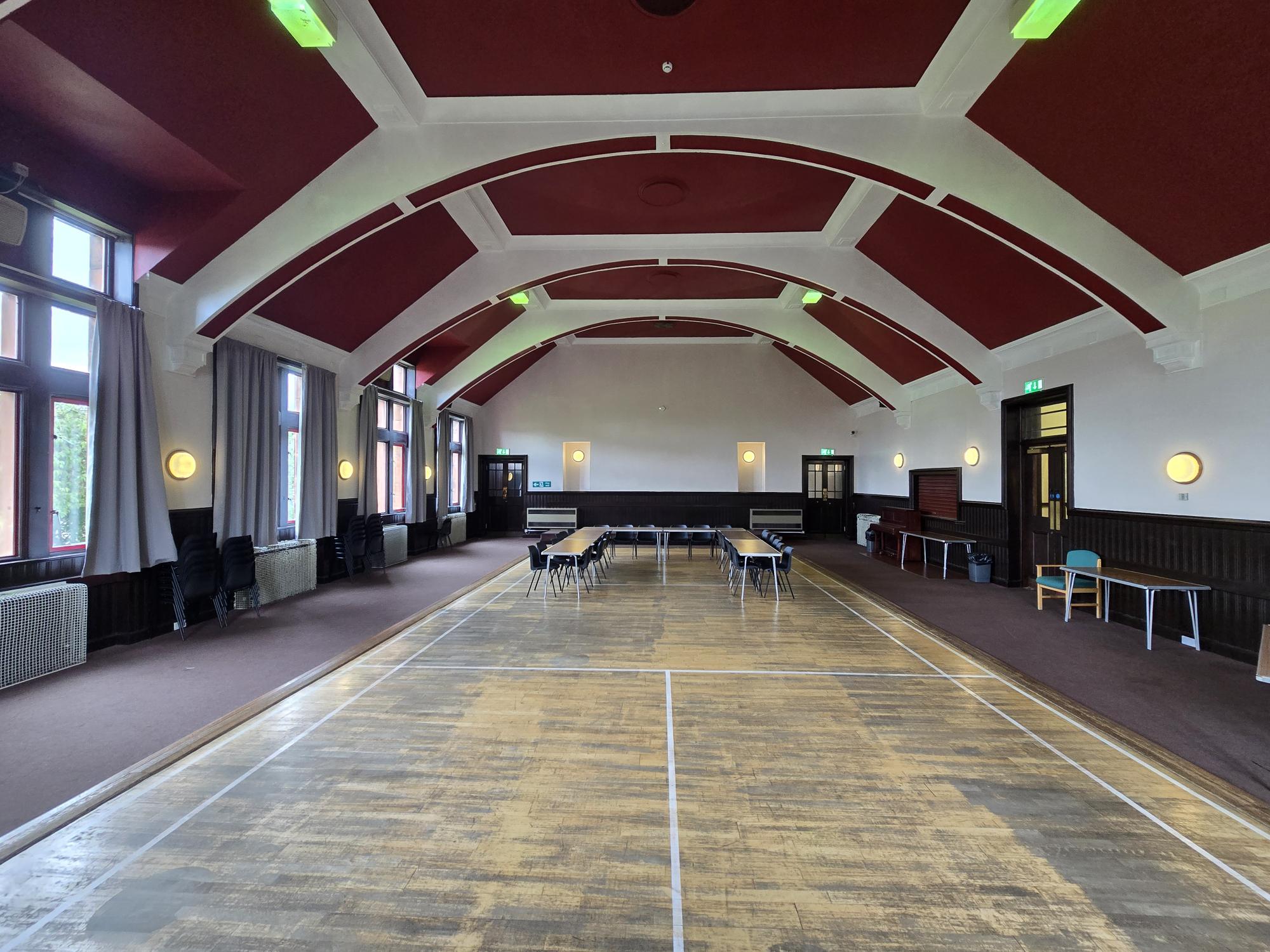 Large hall with arched red-and-white ceiling, tall windows, and a worn wooden floor marked for sports, with tables and chairs along the sides