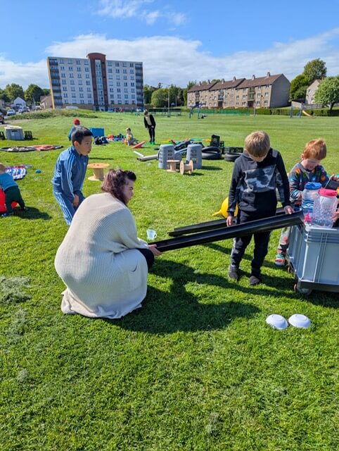 Group of children and an adult using black tubes for a play activity on a grassy field.