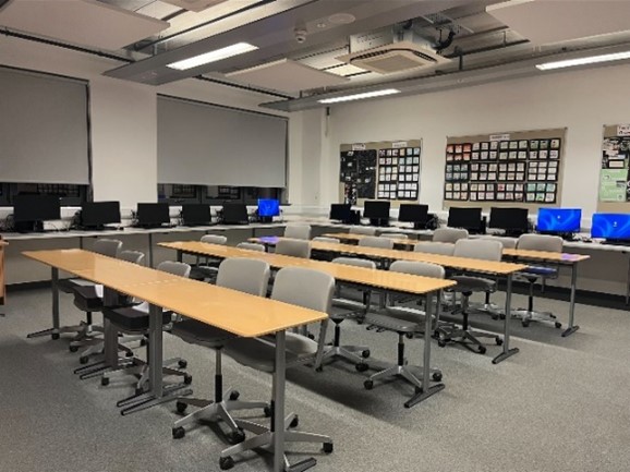 a computer lab with rows of desks, each equipped with a computer monitor. A projector is mounted on the ceiling