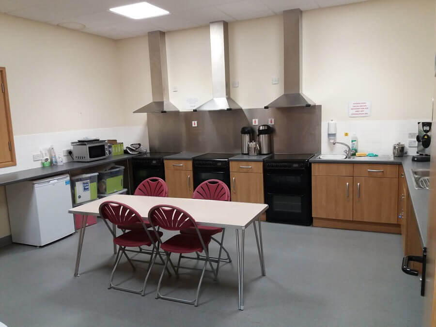 A clean, modern kitchen. It has multiple ovens and hoods. A table with red chairs sits centrally