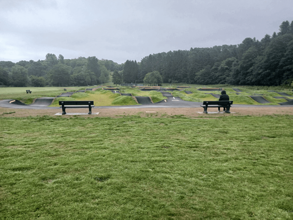 Cycle track within grassy field, benches, and forested background under an overcast sky