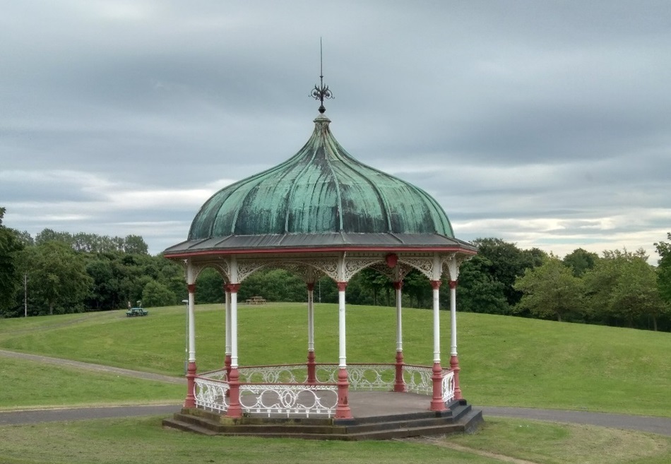 Bandstand with green dome style roof and a red and white pillars in a grassy park and trees in the background