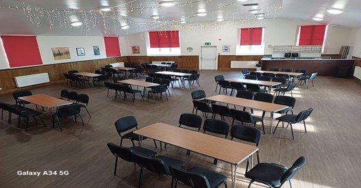 A large community hall set up with numerous rectangular tables and black chairs, featuring wood-effect flooring, half-timbered walls, and bright red window blinds.