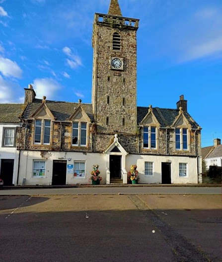 A historic stone building with a tall square clock tower and spire, featuring white-painted lower walls and several gabled windows.