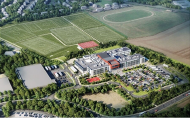 An aerial view of a large secondary school campus surrounded by greenery. In the centre is a modern, low‑rise school building with light‑coloured walls and a rectangular layout, set among car parks, footpaths, and landscaped areas. Behind the school are multiple outdoor sports pitches marked with clear white lines, including several football pitches arranged in a grid. Trees and residential buildings border the site, with open fields visible beyond the sports area, giving a clear sense of scale and the school’s extensive outdoor facilities