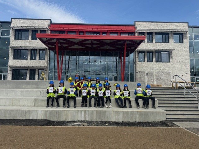 Group of pupils wearing high-visibility vests and blue safety helmets sit in a row on concrete steps outside a modern building with large glass panels and a red-framed entrance canopy. Each person holds a white sign with a single letter, together spelling “100 DAYS TO GO.” The background shows the building façade with light stone walls and windows under a partly cloudy blue sky.