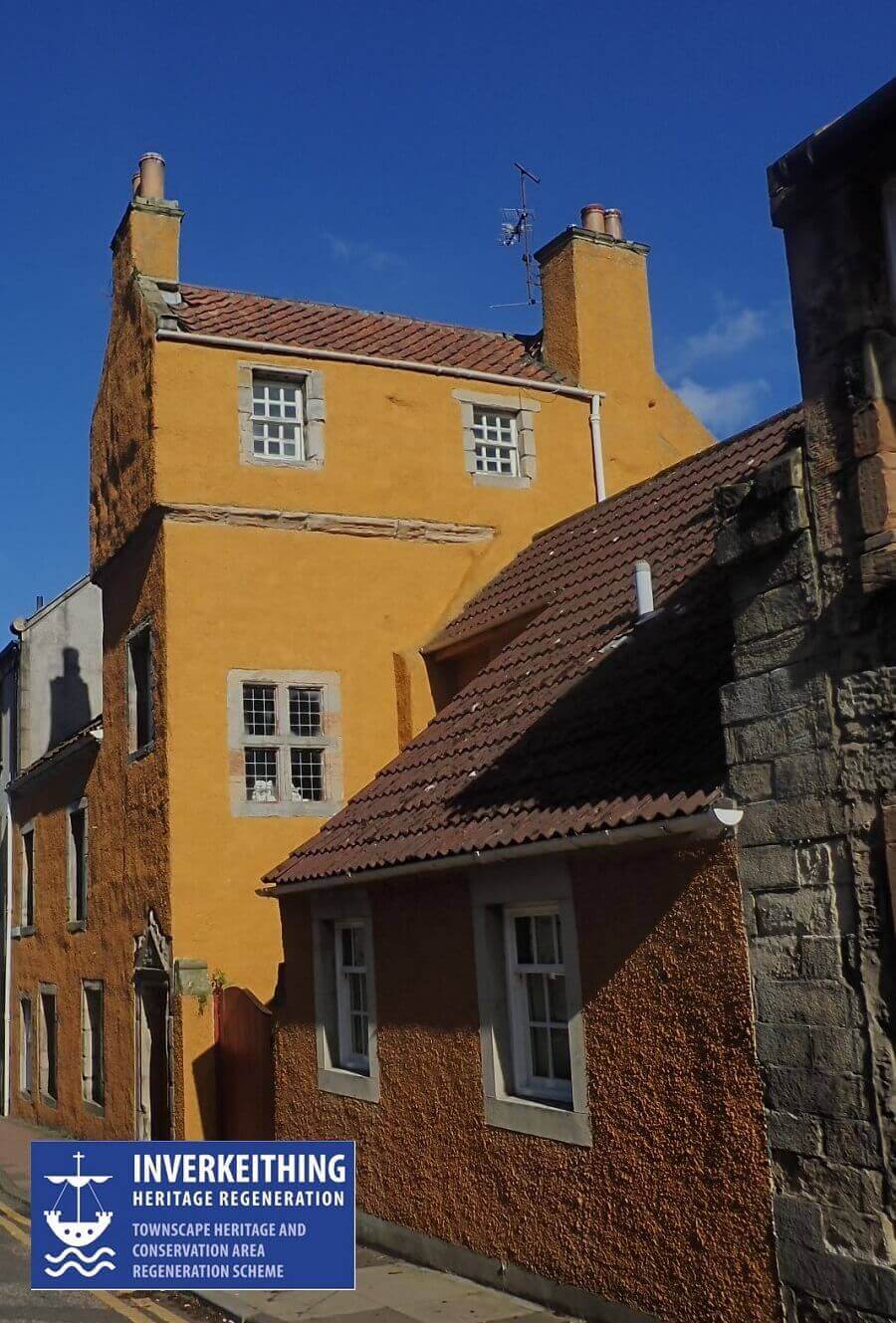 A photograph of a tall, historic building with textured orange‑ochre walls and red‑tiled roofs, situated on a narrow street. The building has multiple levels with small, white‑framed windows and a chimney at the top. Bright sunlight casts strong shadows across the façade. In the bottom left corner, a blue logo reads: ‘Inverkeithing Heritage Regeneration – Townscape Heritage and Conservation Area Regeneration Scheme.