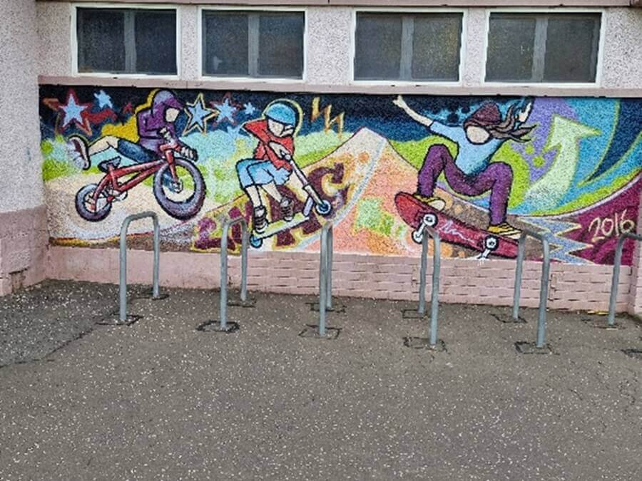 bike rack A vibrant outdoor mural of a cyclist, scooter, and skateboarder. Bike racks stand in front.