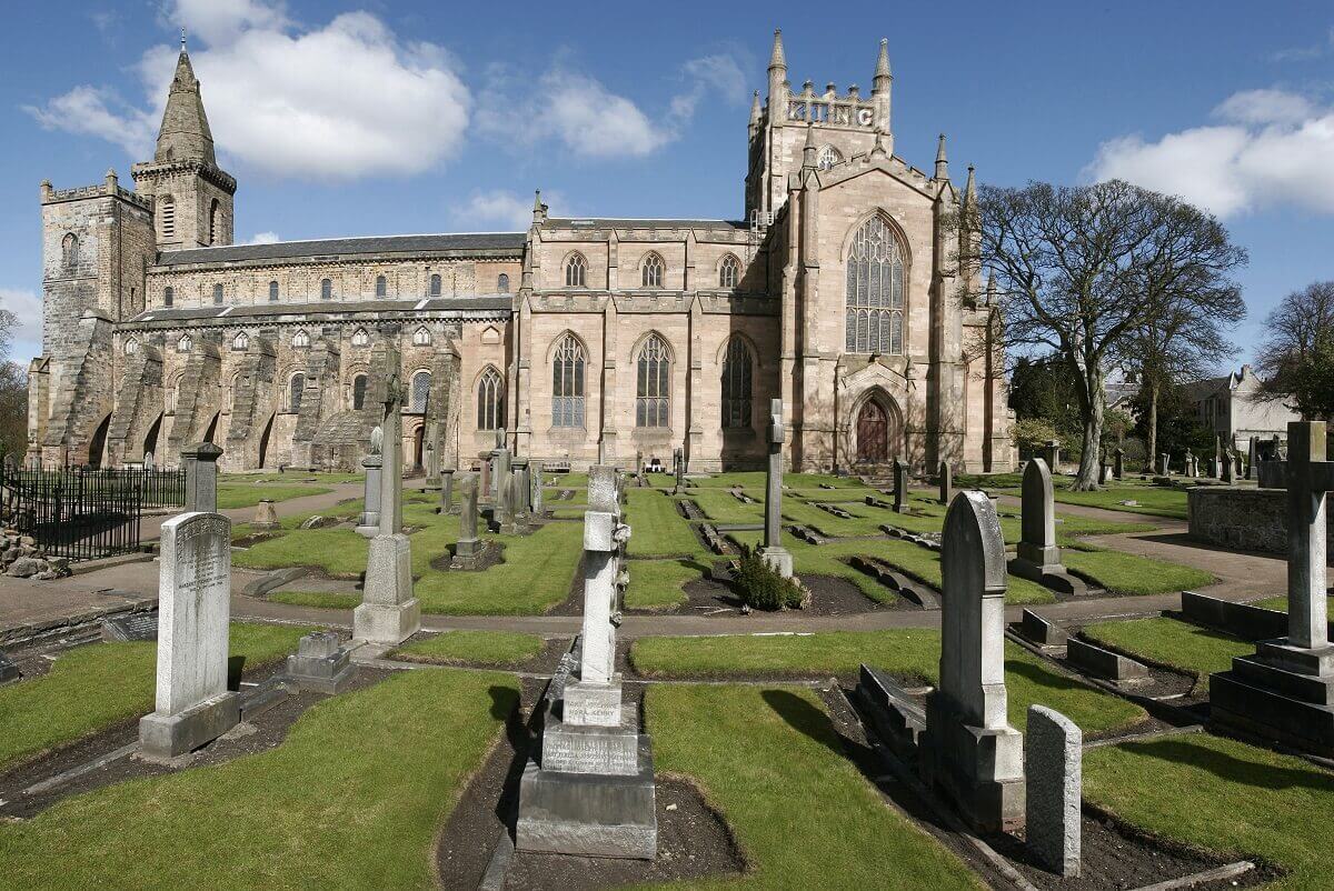 A wide view of a historic stone church with large arched windows, a square tower on the left side, and detailed Gothic-style architecture. In the foreground is a graveyard with rows of old headstones and neatly cut grass. The sky above is bright blue with scattered white clouds, and several mature trees stand near the church.