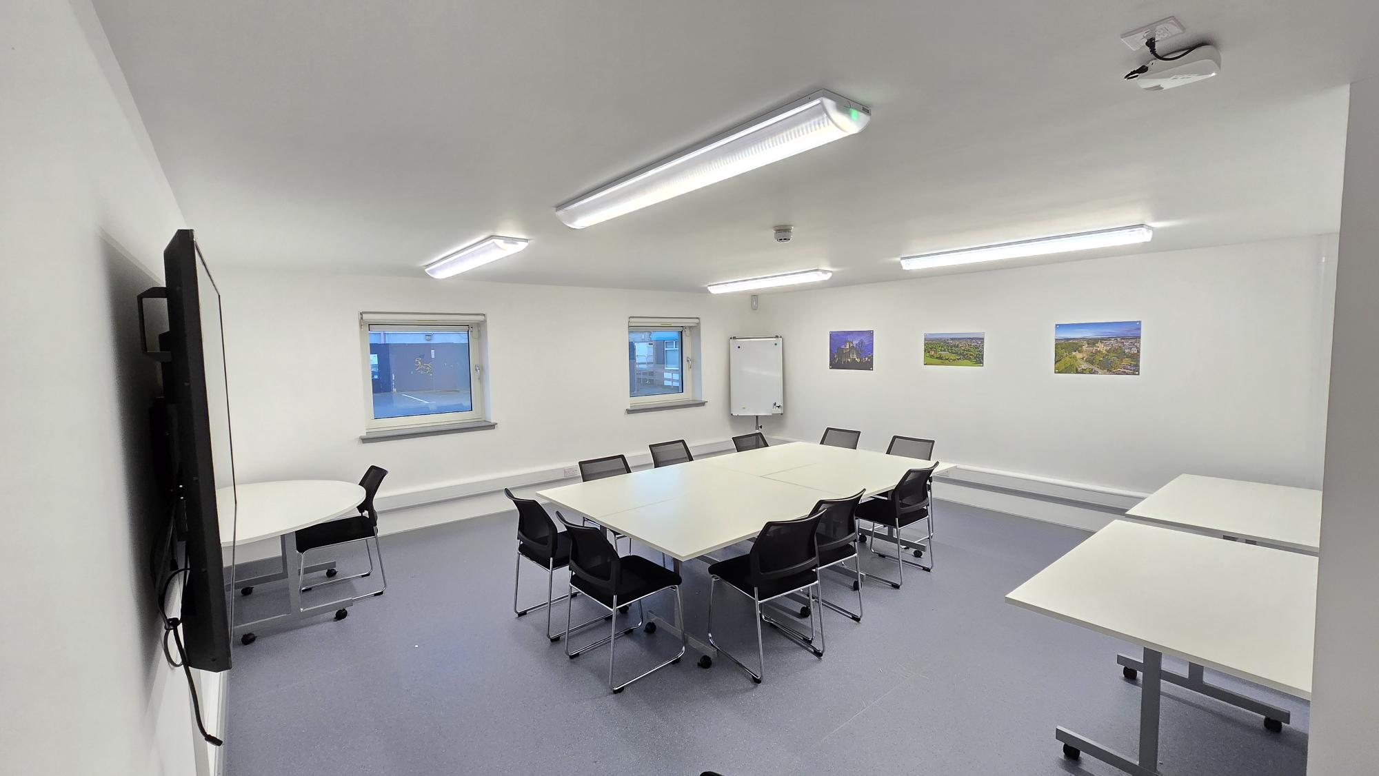 Abbeyview Hub Training & Meeting Space Meeting room with white walls, tables arranged together with chairs, a wall-mounted screen, and a whiteboard, with windows along one side.