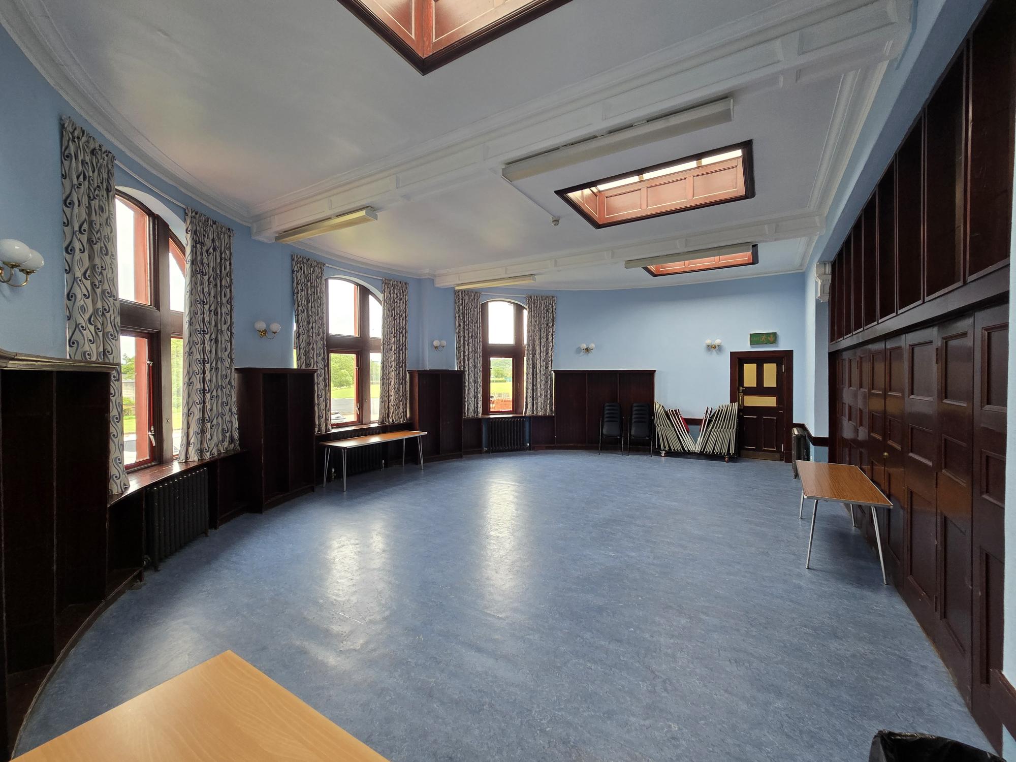 Light blue room with large arched windows, wood paneling, skylights, and tables and chairs stacked along the walls