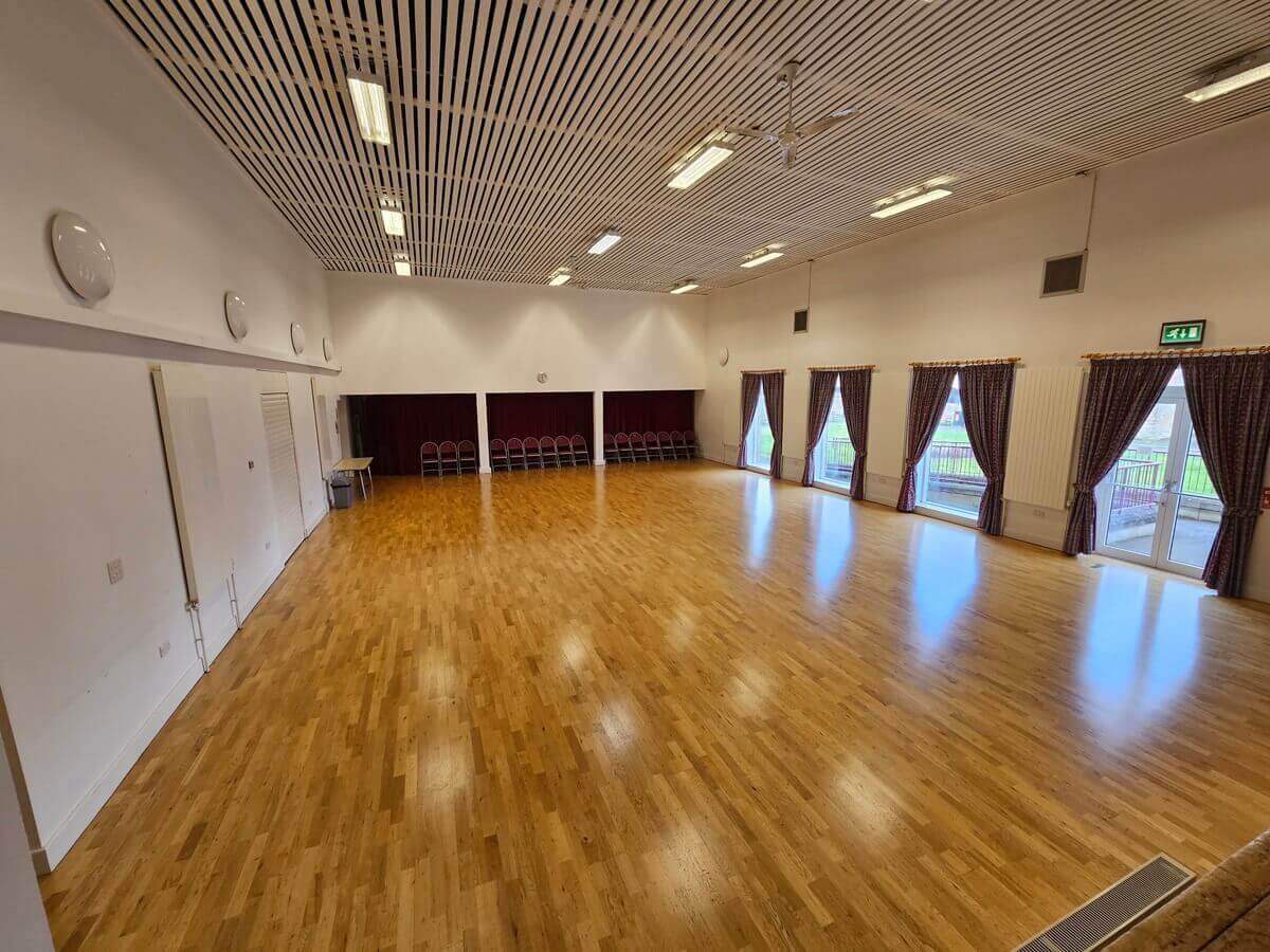 Large hall with a polished wooden floor, high slatted ceiling, stage area with red curtains, and full-height windows with patterned drapes along one side.