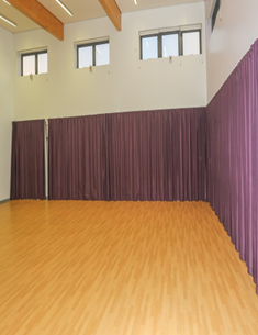 a dance studio with a light wooden floor and white walls. Purple curtains hang along one wall, partially obscuring the view behind them. There are several windows near the ceiling, allowing natural light into the room. Wooden beams are visible on the ceiling.