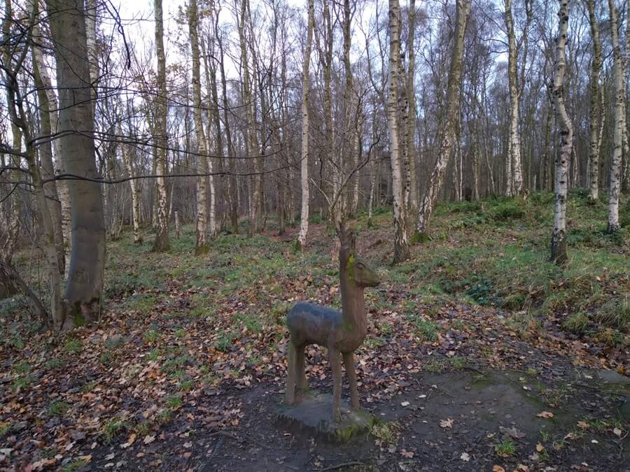 A small deer sculpture stands on a dirt path in a woodland area, surrounded by tall, leafless birch trees and scattered fallen leaves.