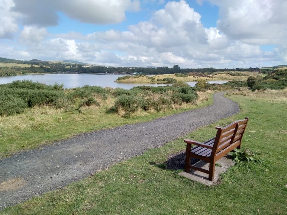 Scenic view at Lochore Meadows Country Park with a bench beside a path looking onto the loch, surrounded by greenery and hills under a partly cloudy sky