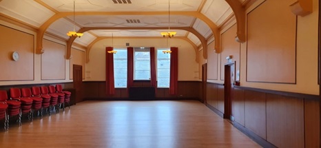 A spacious room with arched ceilings, wood flooring, and a row of stacked red chairs against a panelled wall