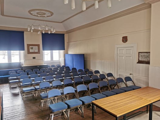 A room filled with rows of blue folding chairs facing a wooden table, featuring white-panelled lower walls, blue window blinds, and two circular chandeliers hanging from a high ceiling.
