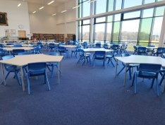 A bright, expansive library or study area featuring blue carpeting and multiple light-colored, uniquely shaped group tables surrounded by blue plastic chairs. Massive floor-to-ceiling windows line the right wall, looking out onto a green landscape. Bookcases and a framed portrait are visible in the background.