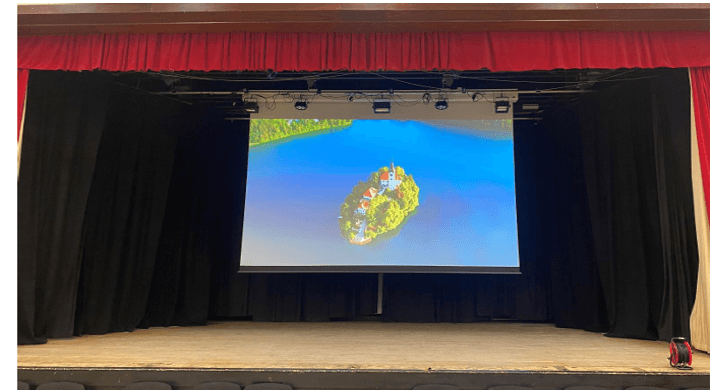 Corn Exchange Projection Screen A theater stage with red and black curtains, featuring a large central projection screen displaying an island in a blue lake.