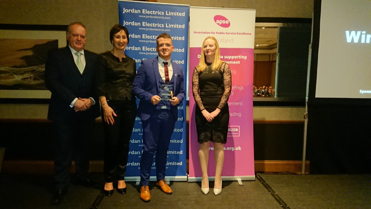 Four people standing in front of banners at the Scottish APSE annual awards event. One person is holding a glass award