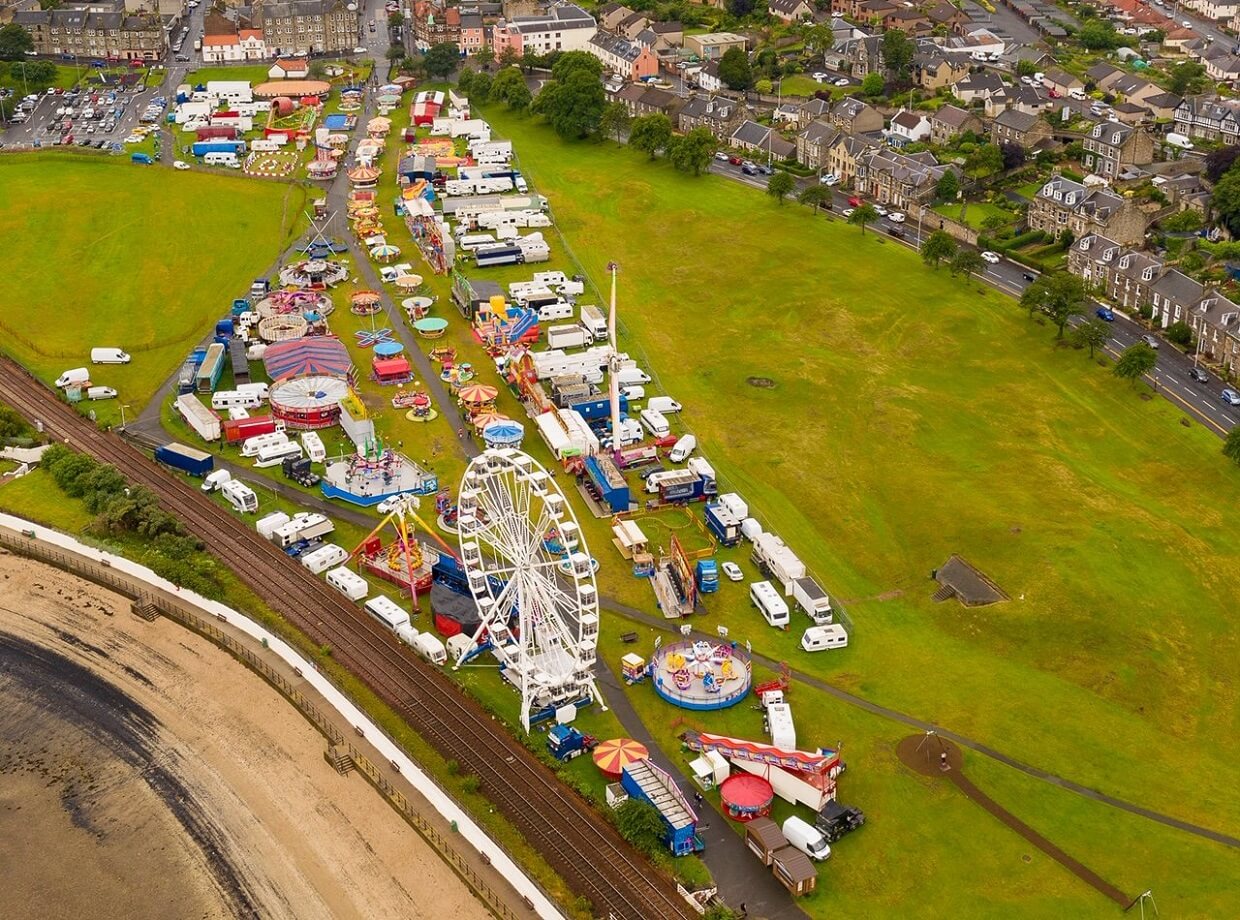 Aerial view of a fairground with rides, stalls, and caravans set up on a large grassy area near a beach and railway tracks, with houses in the background