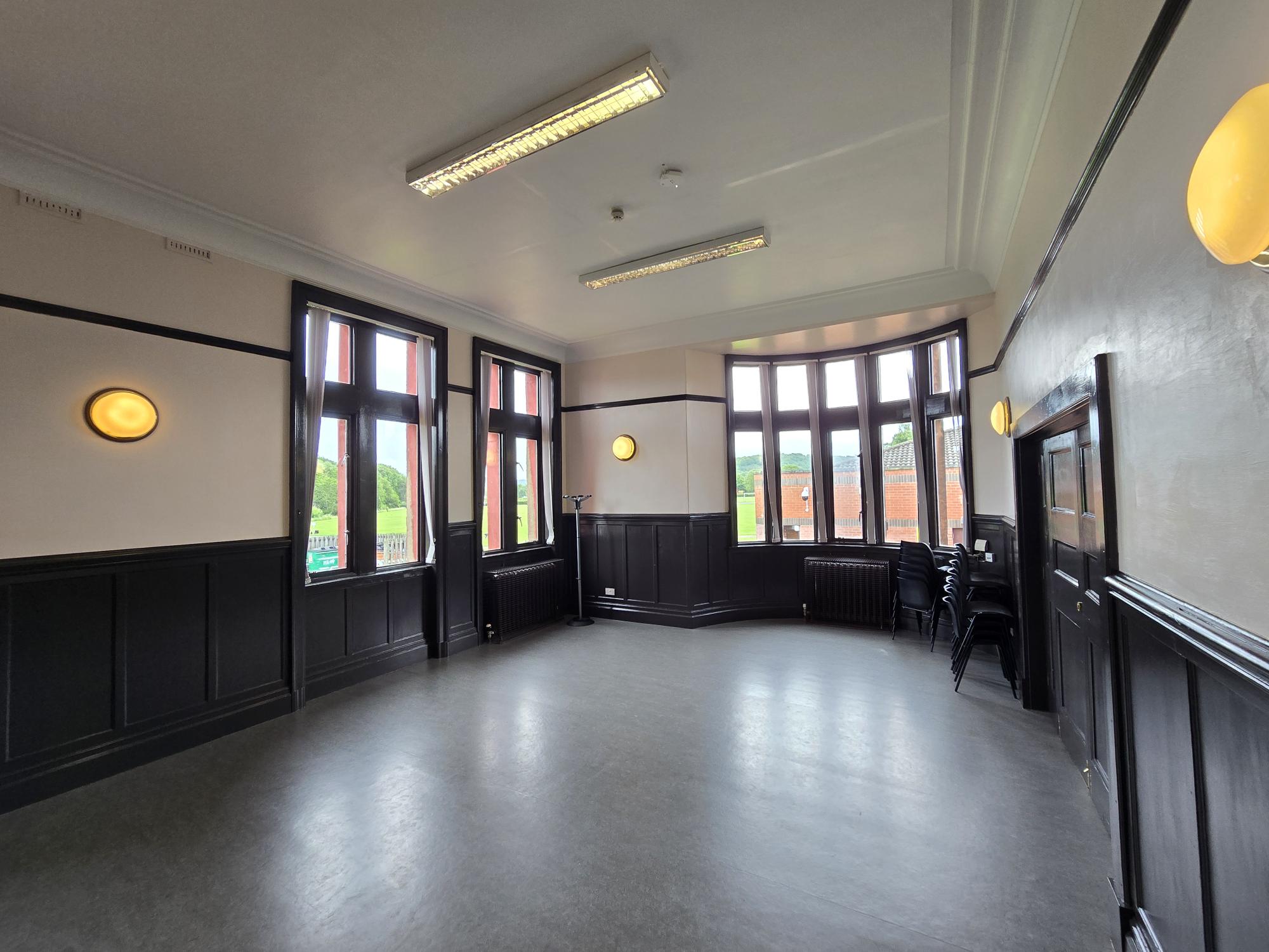 Bright room with tall windows, dark wood panelling, light walls, and stacked chairs in the corner