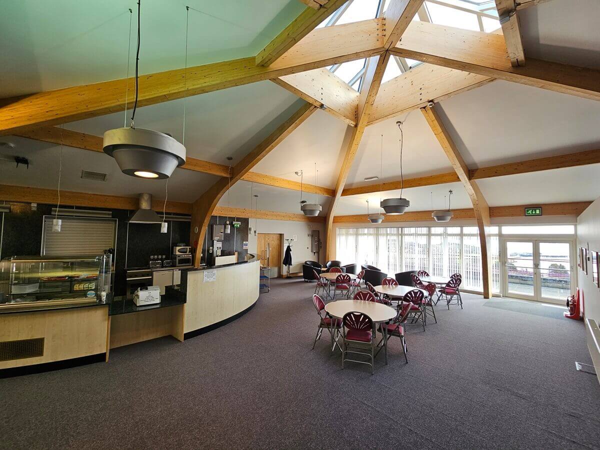 Spacious café-style room with wooden ceiling beams, a serving counter, round tables with red chairs and large windows letting in natural light.