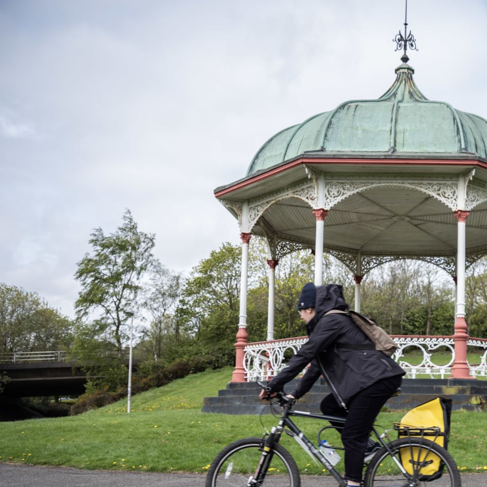 Person riding a bicycle past a bandstand with a green domed roof in a park setting - Active travel gets a boost in Dunfermline 