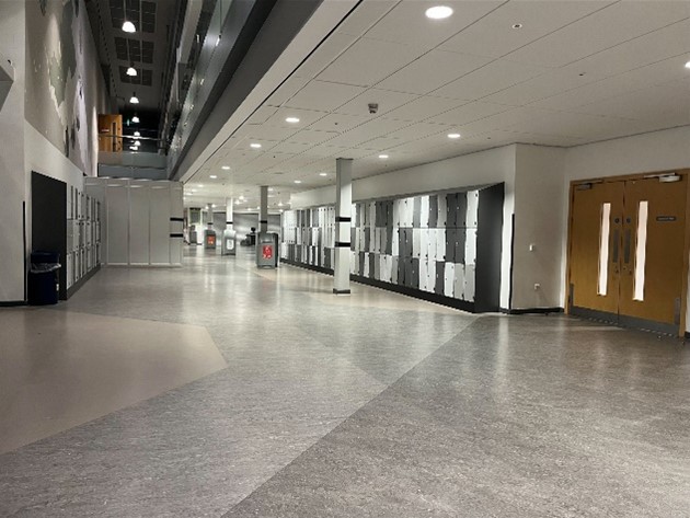  an indoor hallway with a grey speckled floor and a row of lockers along the right wall. The lockers are two-toned, alternating between grey and white. There are double doors at the end of the hallway, and the walls are white