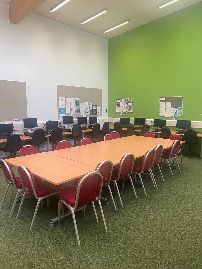  a classroom with a large rectangular table surrounded by red chairs in the foreground. In the background, there are rows of computer desks with monitors and chairs. The walls are painted in white and a light green color, and there are notice boards with papers displayed on them. The floor is covered with green carpet