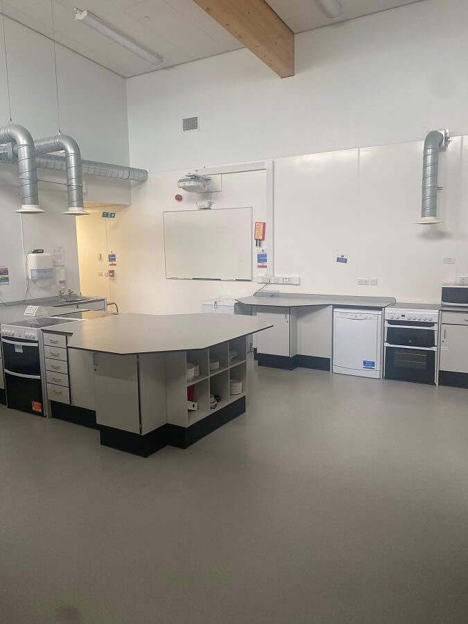 a well-equipped kitchen classroom. There is a central island with a light-colored countertop and storage underneath. Along the walls, there are various appliances including ovens, a dishwasher, and sinks, all with stainless steel ventilation hoods above. A whiteboard is visible on the back wall. The floor is a light grey.