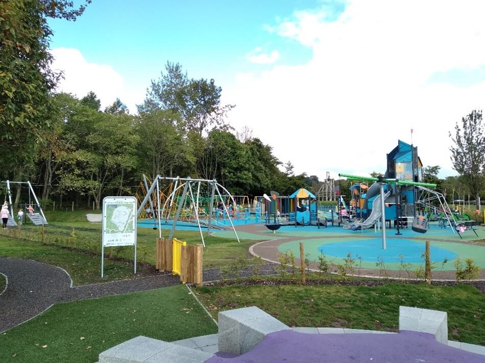 Playground with swings, slides, and climbing structures, surrounded by trees and walking paths.