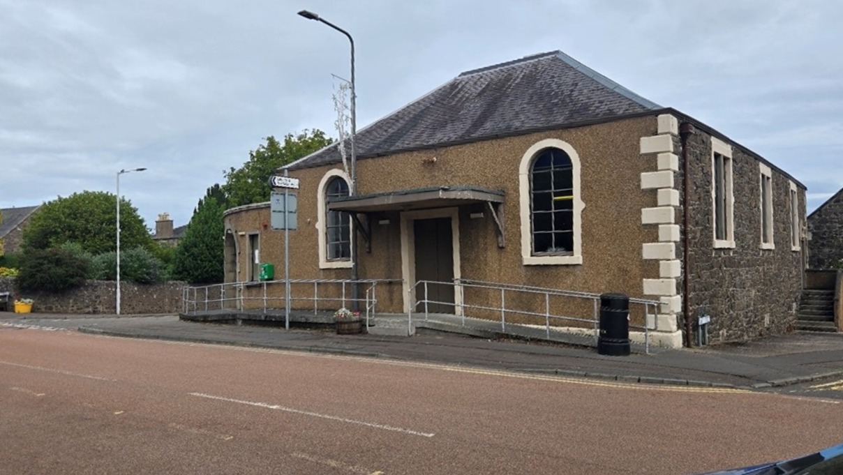 A single-story tan and stone building with arched windows and a ramped entrance under a small canopy.